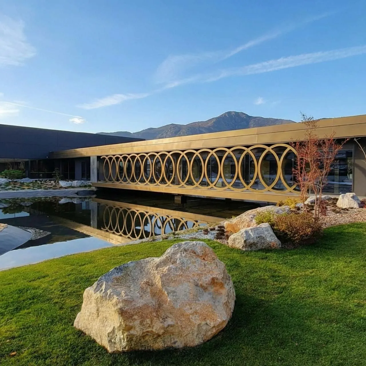 Bernardi's factory in Italy: a long building with a facade of linked gold rings over a reflecting pool, a lawn and a boulder in front and mountains behind.