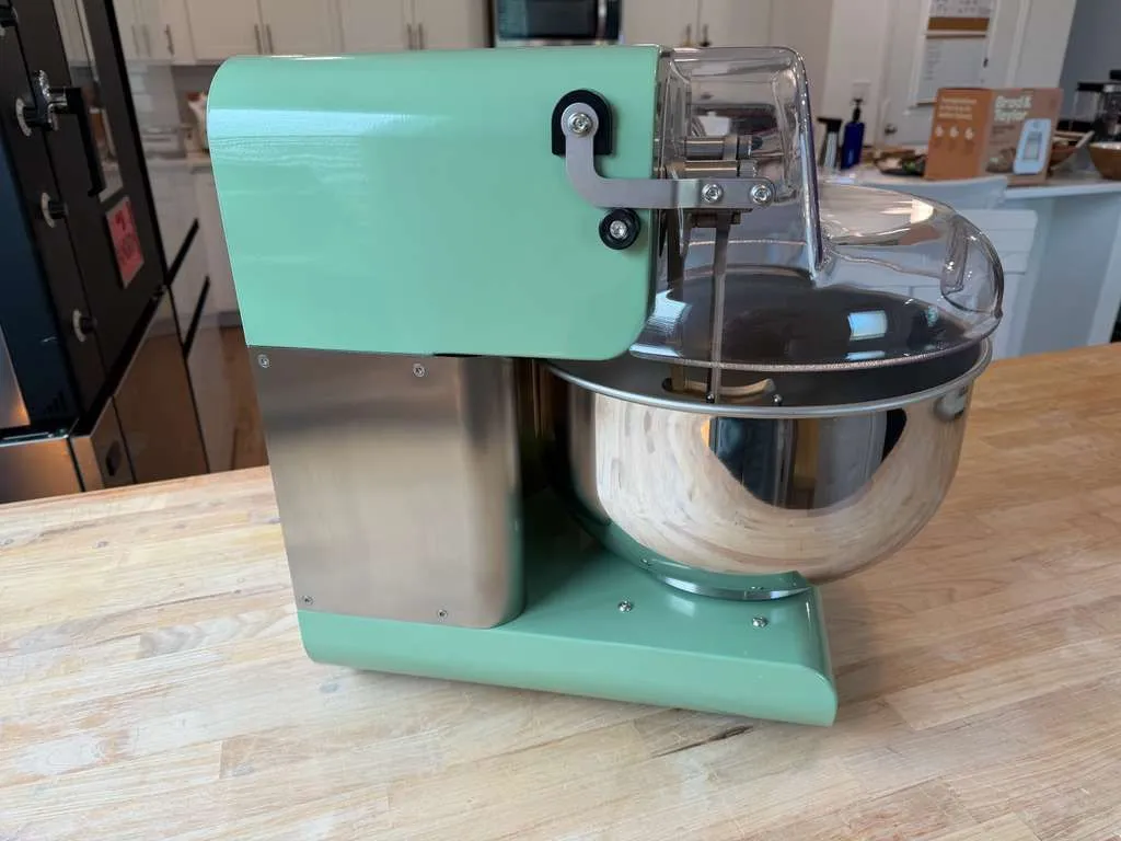 A green My Miss Baker from the side: the clear safety cover on its hinge over the steel bowl.